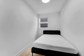 A narrow room with a single bed, a window with blinds, and a light fixture on the ceiling. - Newly Renovated Apartment in Chinatown (Ottawa)