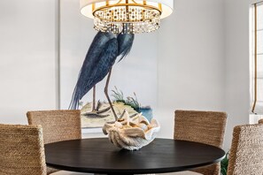 Une table de salle à manger ronde, entourée de chaises en osier, un lustre et un tableau représentant un oiseau, accroché au mur.