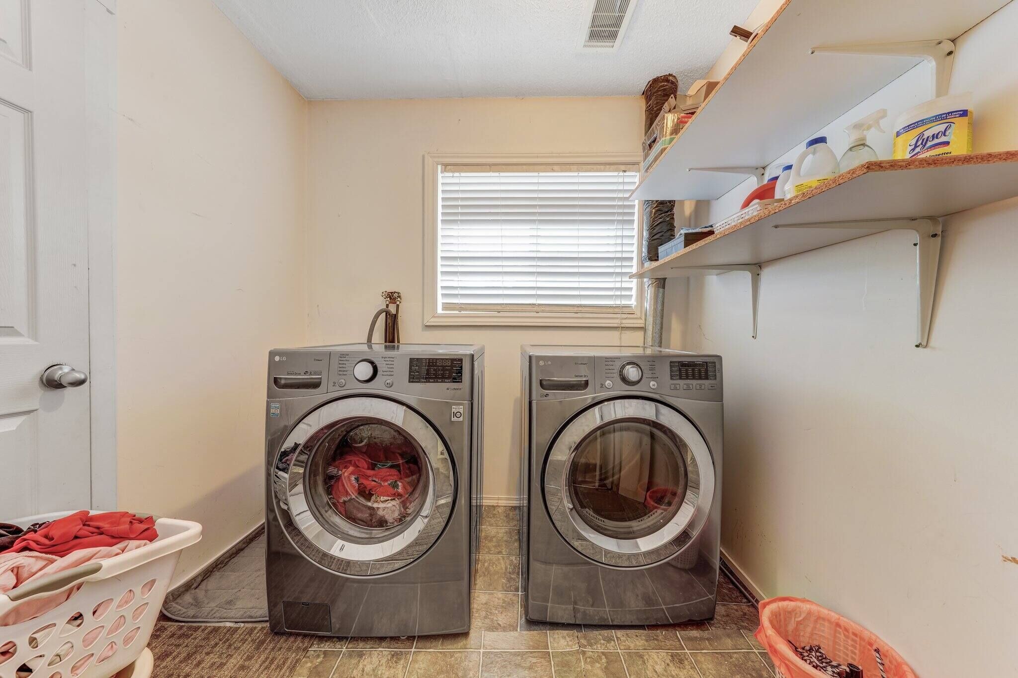 A laundry room with two front-loading washing machines, a window with blinds, a shelf with cleaning supplies, and a laundry basket.