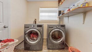 Un cuarto de lavandería con dos lavadoras de carga frontal, una ventana con persianas, un estante con productos de limpieza y una cesta de ropa.