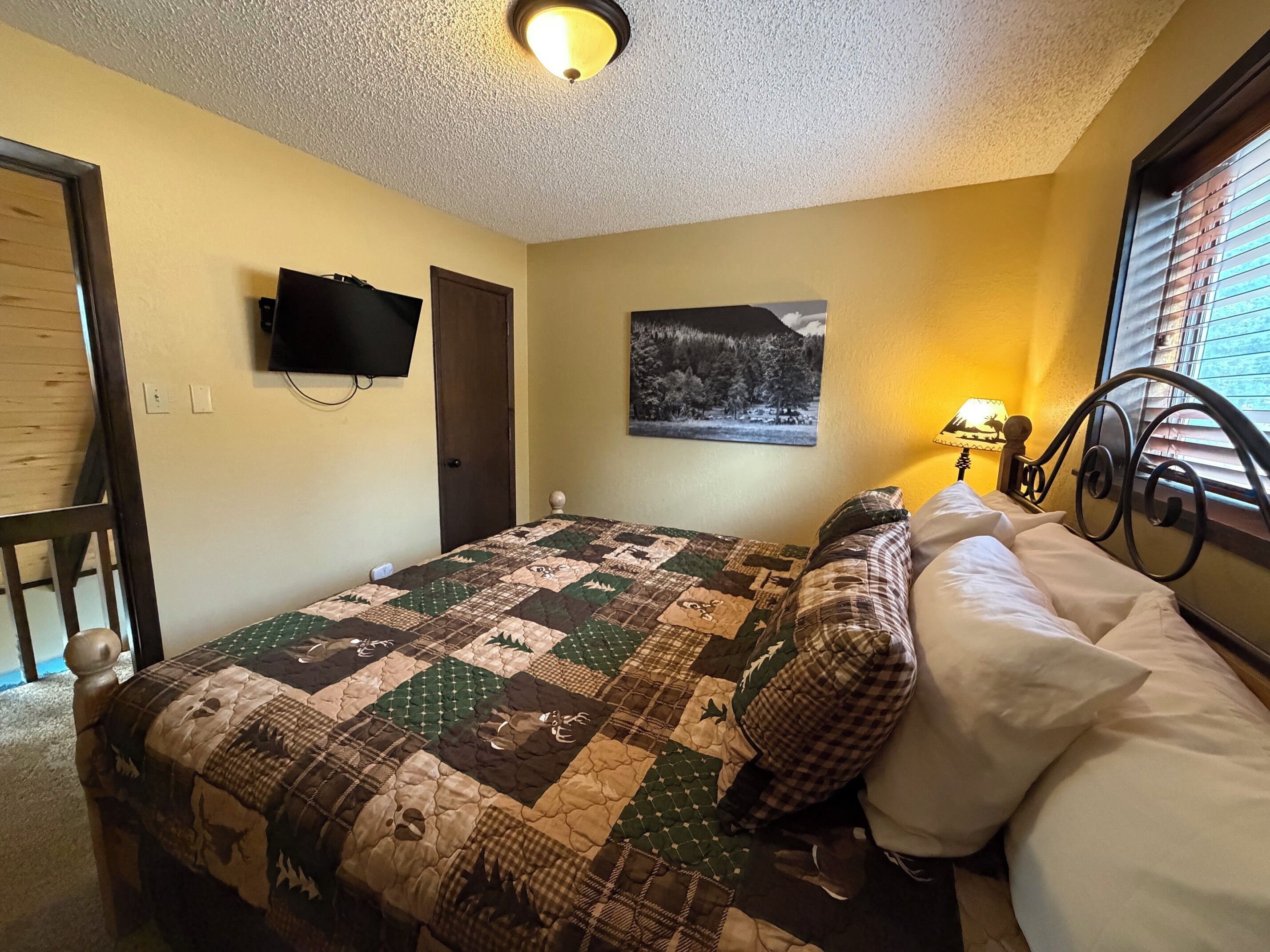 A bedroom with a bed, a television mounted on the wall, a door, and a window with blinds.