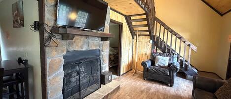 A cozy living room with a stone fireplace, a flat-screen TV mounted above it, and a wooden staircase leading to an upper level.