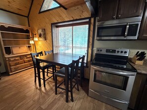 Een keuken met een houten tafel, stoelen, een fornuis, een magnetron en een raam met jaloezieën.