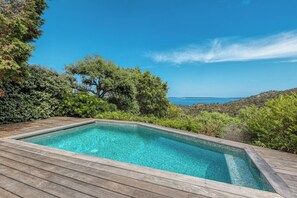 A rectangular swimming pool with clear blue water, surrounded by a wooden deck, set against a backdrop of lush greenery and a distant view of the sea. - Two-bedroom apartment with ocean views in Gaou Bénat—pool, private estate, beaches 5 minutes away (Bormes-les-Mimosas)