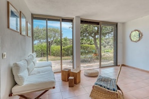 A modern room with a sofa, a small wooden table, and a woven chair. Large glass doors lead to an outdoor area with a bench and trees. - Two-bedroom apartment with ocean views in Gaou Bénat—pool, private estate, beaches 5 minutes away (Bormes-les-Mimosas)
