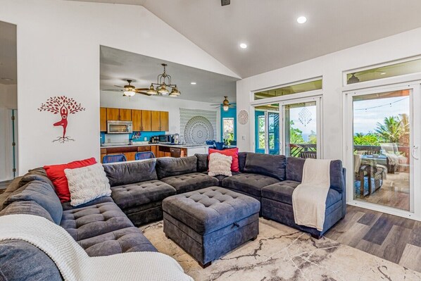 A spacious living area with a large sectional sofa, a dining area with a kitchen in the background, and a sliding glass door leading to an outdoor patio.
