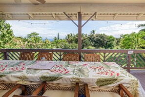 A covered outdoor dining area with a table set for two, overlooking a lush landscape with palm trees and a view of the ocean.