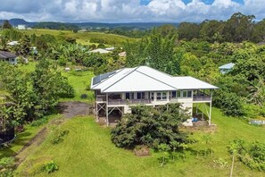 A large house with a spacious deck and multiple windows, surrounded by lush greenery and a hilly landscape.
