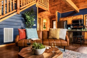 A cozy cabin interior with a wooden staircase, a living area with a leather sofa, a dining table with a plant, and a kitchen with wooden cabinets. - Quaint Cute Cabin|Pool Table|4min to Pkway|New|Hot Tub (Pigeon Forge)