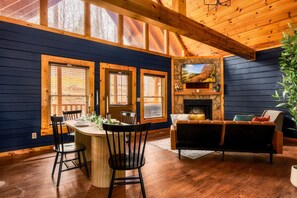 A dining area with a wooden table and chairs, large windows, a stone fireplace, and a view of trees. - Quaint Cute Cabin|Pool Table|4min to Pkway|New|Hot Tub (Pigeon Forge)