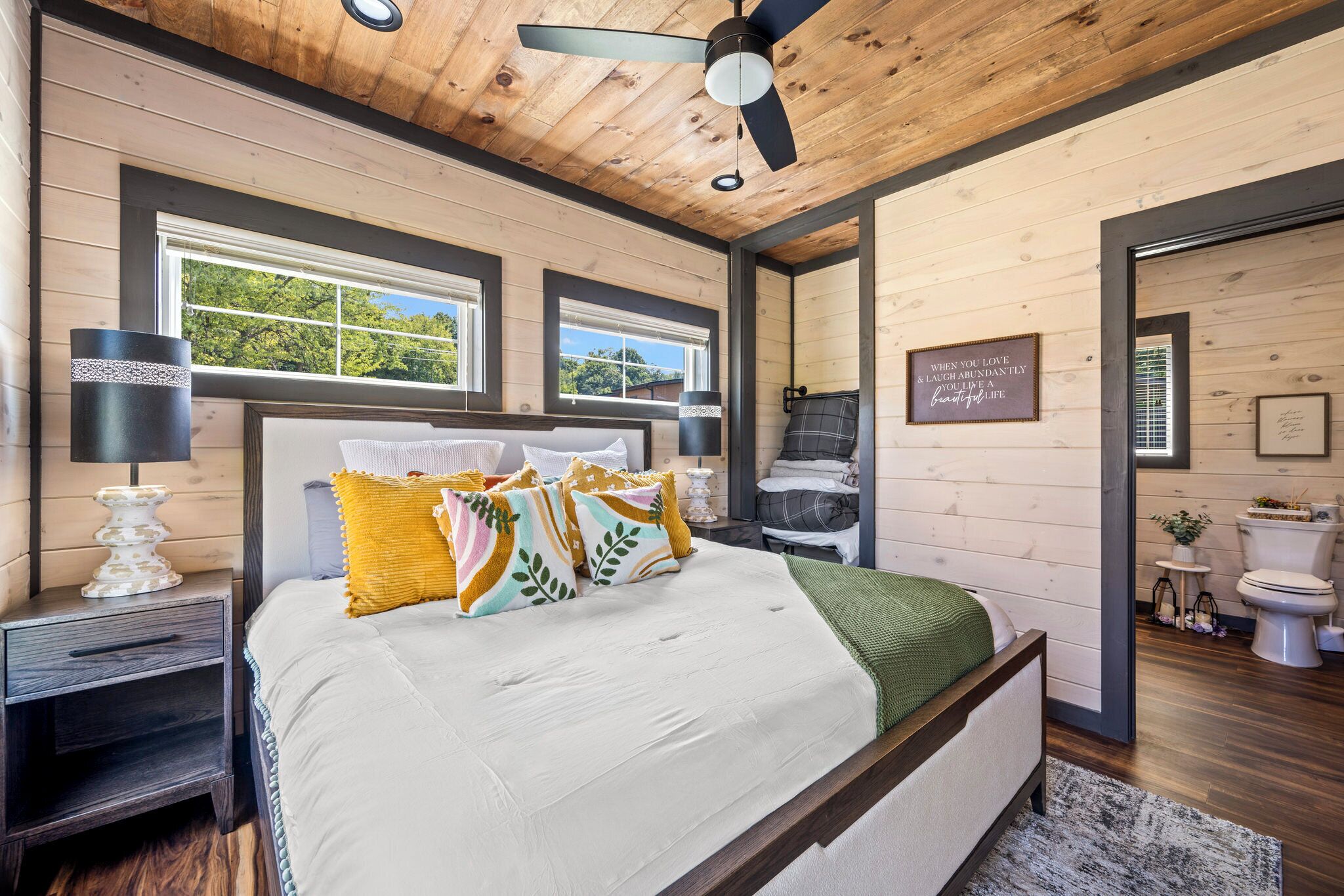 A bedroom with a bed, nightstands, a ceiling fan, windows, and a bathroom with a toilet and sink.