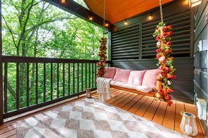 A wooden deck with a hanging swing, a cushioned bench, and a view of greenery. - Romantic Cabin w/ Hot Tub • 3mi to Pigeon Forge • King Bed • Fireplace (Pigeon For)