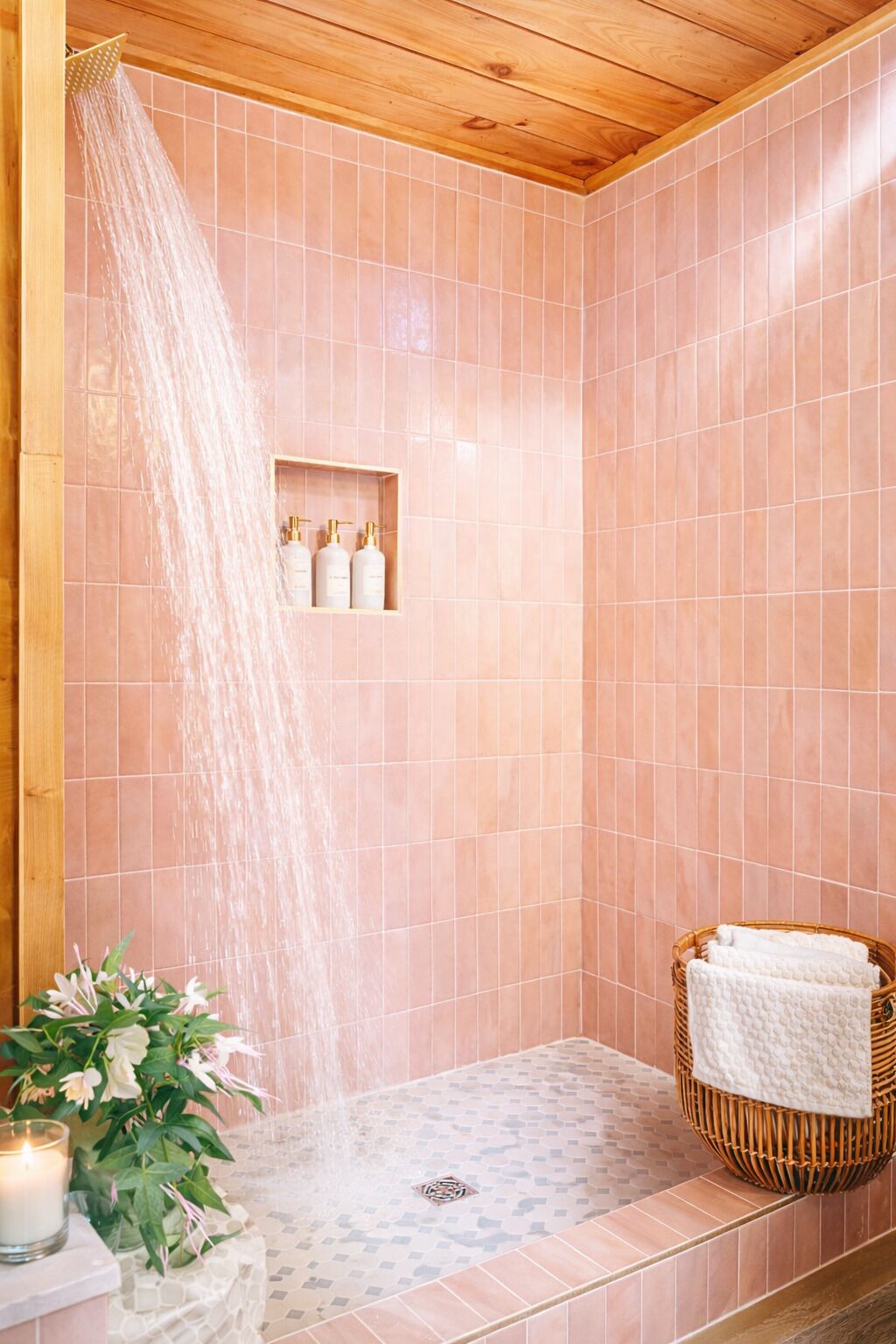 A shower with a wooden ceiling, pink tiles, a shelf with toiletries, a candle, and a wicker basket with towels.