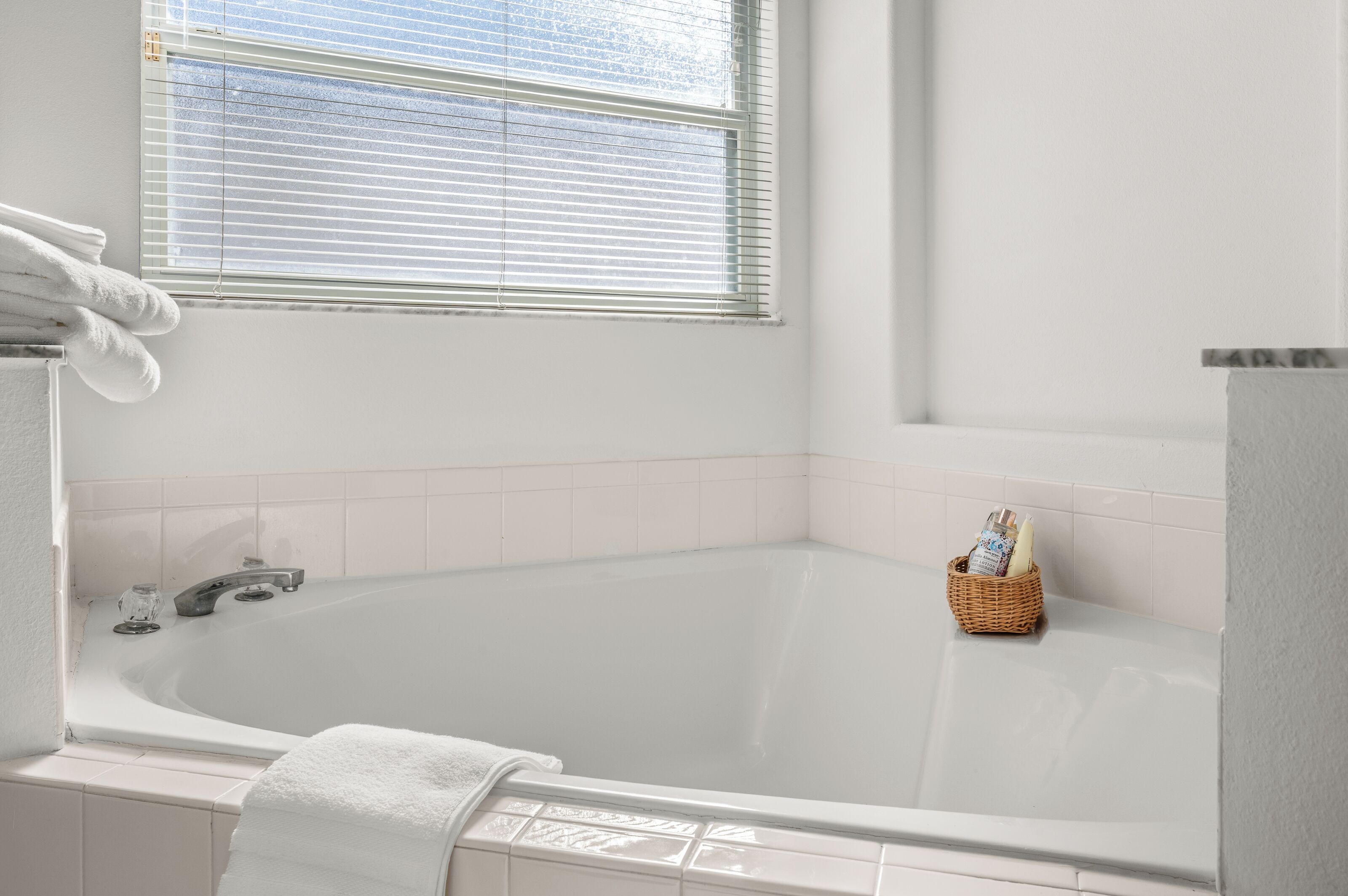 Een badkamer met een witte badkuip, een raam met zonwering en een klein rieten mandje met toiletartikelen.