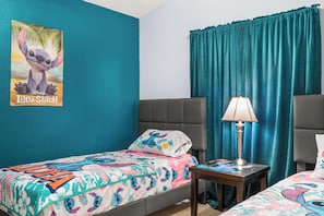 Een hotelkamer met een blauwgroene wand, een poster van Lilo & Stitch, twee bedden met kleurrijk beddengoed, een nachtkastje met een lamp en een grijze hoofdeinde.