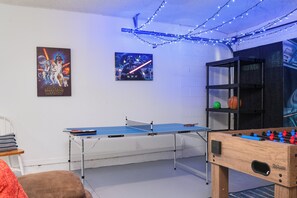 Een kamer met een tafeltennistafel, een tafelvoetbaltafel en twee posters aan de muur.