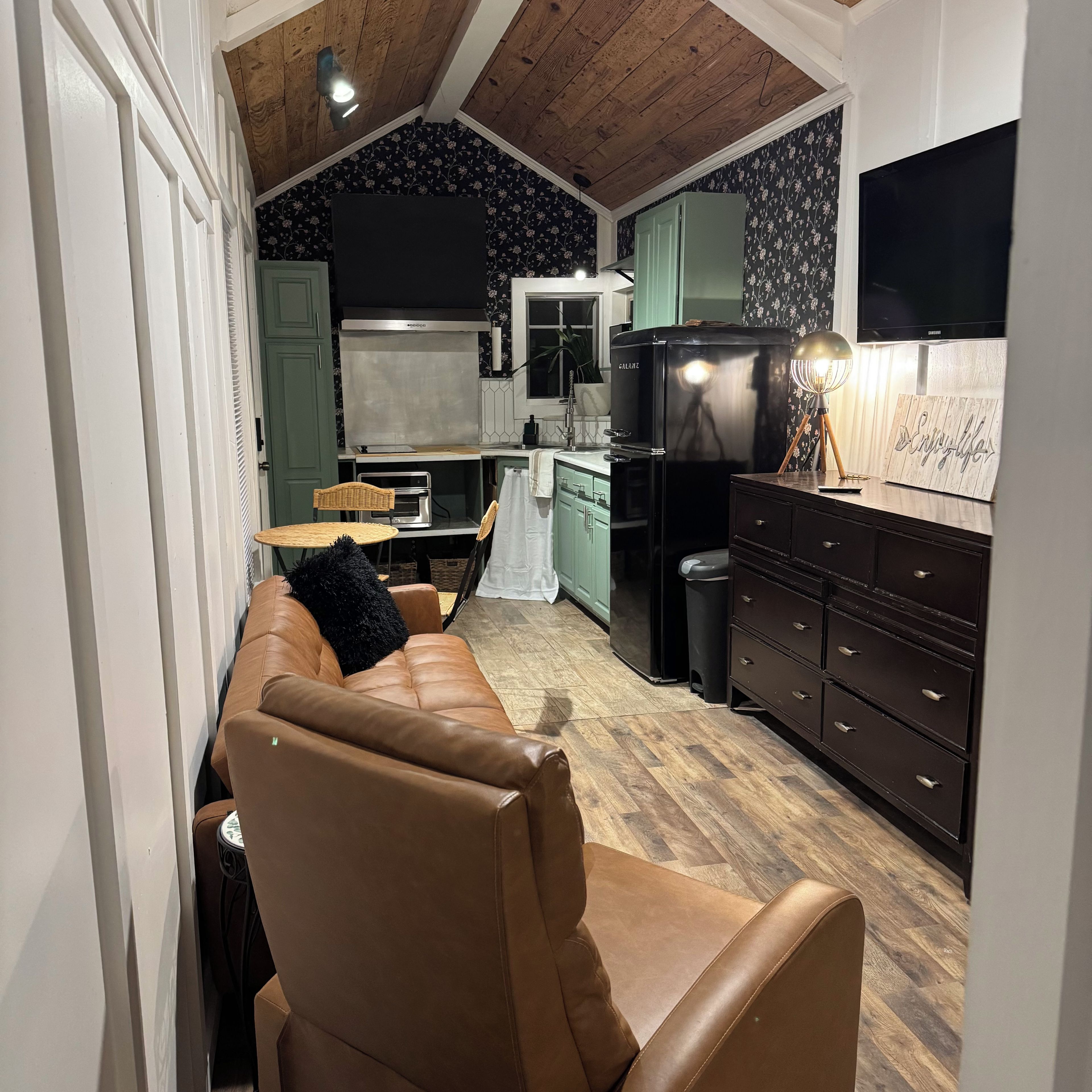 A cozy living room with a leather sofa, a dining table, and a kitchen area with a refrigerator and a television.