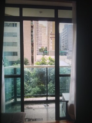 Ein Zimmer mit großem Fenster, einem Balkon und Blick auf hohe Gebäude und Grünflächen.