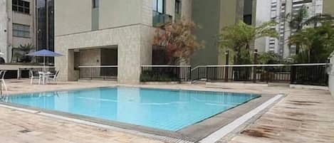 A swimming pool with lounge chairs and a table, surrounded by a fence and buildings.