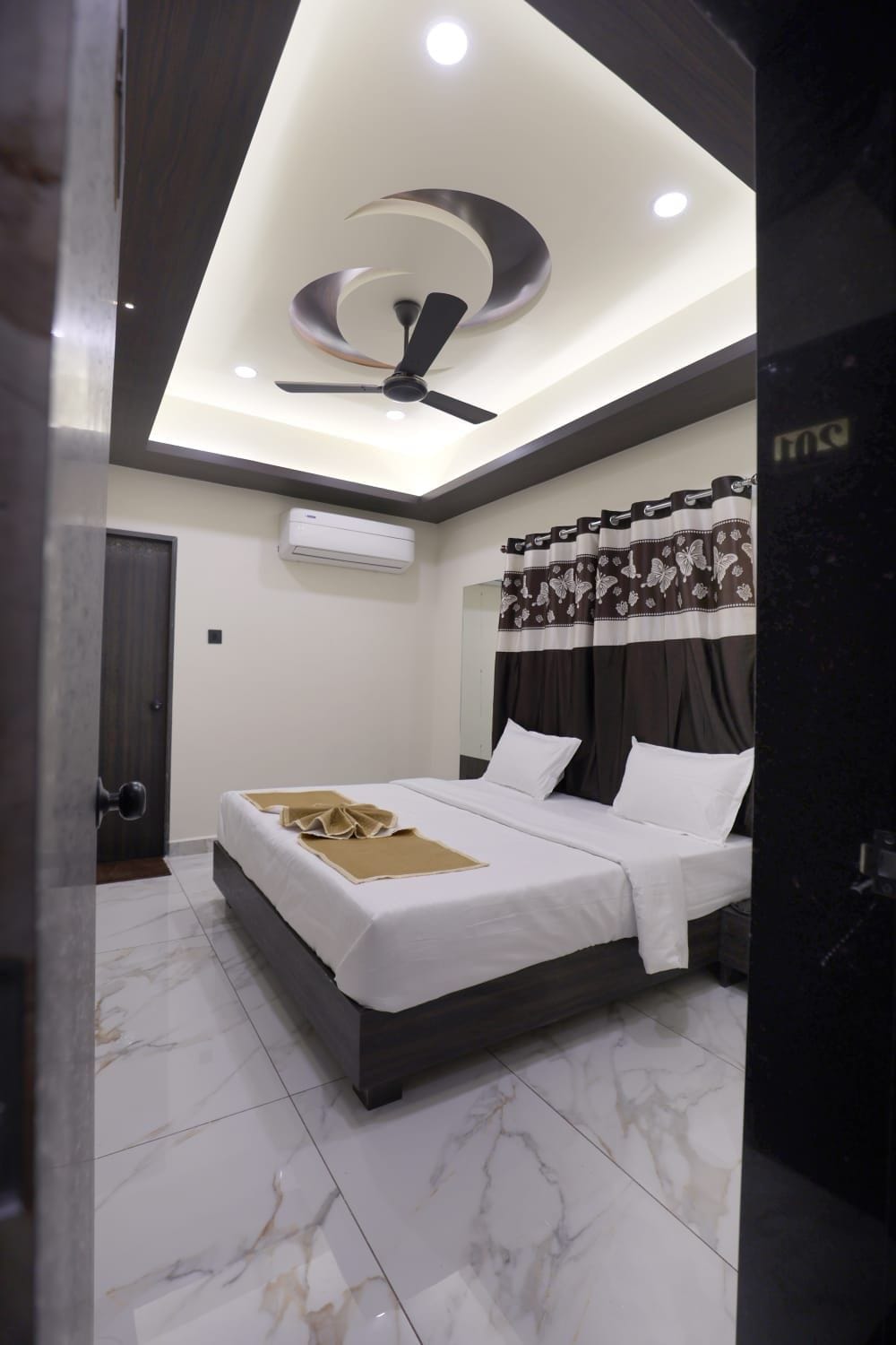 A modern bedroom with a bed, ceiling fan, air conditioning unit, and patterned curtains.