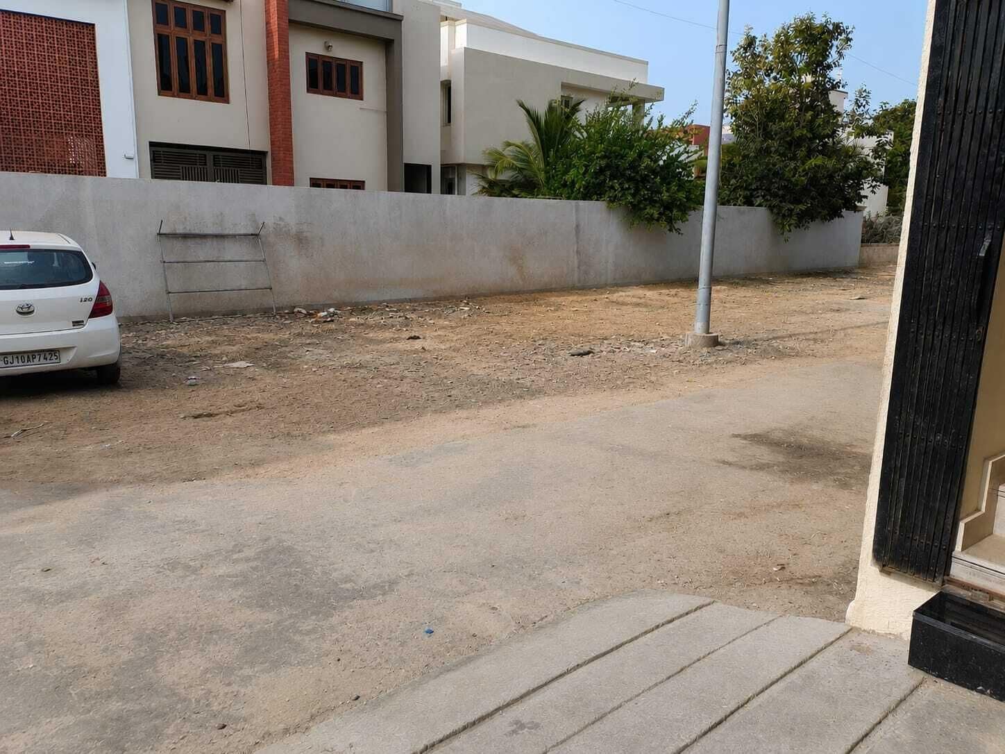 Une voiture blanche est garée sur un chemin de terre, avec un mur en béton et un bâtiment en arrière-plan.