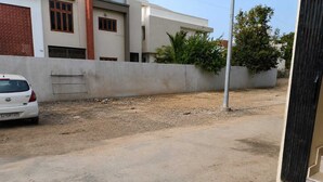 Une voiture blanche est garée sur un chemin de terre, avec un mur en béton et un bâtiment en arrière-plan.