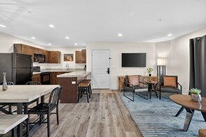 A modern kitchen with wooden cabinets, a refrigerator, and a microwave. There is a dining area with a table and chairs, and a living room with a sofa and a TV. - 4-A Arches Redcliff Condo (Ground Floor) (Moab)