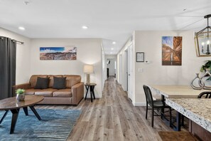A modern living room with a brown leather sofa, a coffee table, and a rug. There is a dining area with a wooden table and chairs. The room has wooden flooring and white walls. - 4-A Arches Redcliff Condo (Ground Floor) (Moab)