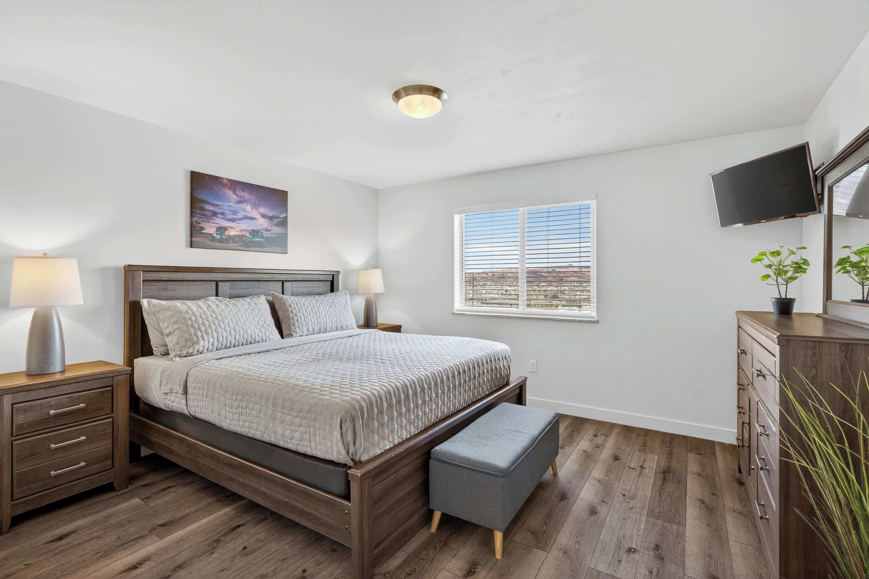 Ein Schlafzimmer mit einem Bett, Nachttischen, einem Fenster mit Jalousien, einer Deckenleuchte und einem an der Wand befestigten Fernseher.