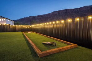 A well-lit outdoor area with artificial lights along a fence, a green artificial turf, and a wooden table with cushions. - Moab Desert Oasis Pool Spa Red Rock Views (Moab)