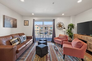 A living room with a brown leather sofa, a red patterned armchair, a black ottoman, a television, and a patterned rug. - 2-E Copper Ridge Condo (Moab)