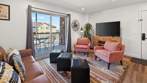 Ein Wohnzimmer mit einer Couch, zwei Sesseln, einem Fernseher und einem Balkon mit Tisch und Stühlen.