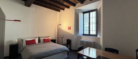 Una habitación con cama, mesa, silla, ventana y radiador.
