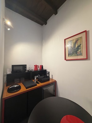 Ein kleiner Büroraum mit Schreibtisch, einer Mikrowelle, einer Kaffeemaschine, einem gerahmten Bild an der Wand und einem schwarzen Stuhl.