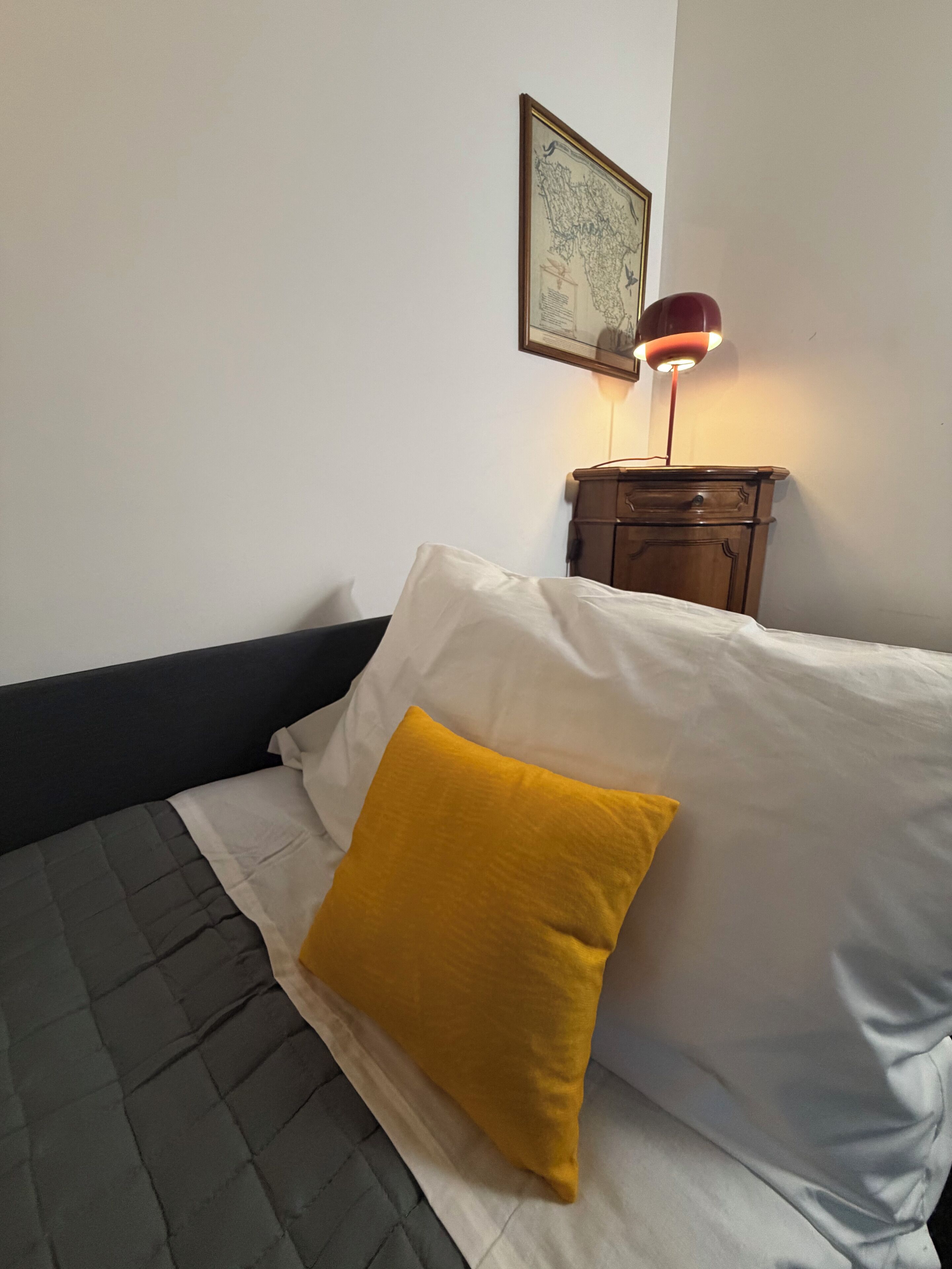 Ein Bett mit einem weiß-gelben Kissen, einem dunklen Kopfteil, einem Nachttisch aus Holz mit einer Lampe und einer gerahmten Karte an der Wand.