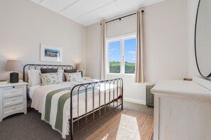 Ein Schlafzimmer mit Bett, Nachttischen, einem Heizkörper, einem Fenster mit Vorhängen und Blick auf Grünflächen.