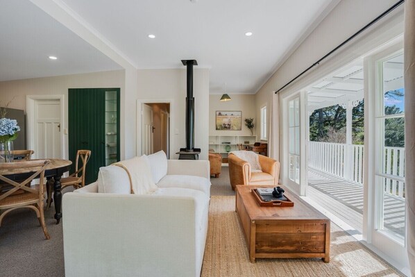 A spacious living room with a fireplace, a dining area with a round table and chairs, and large windows offering a view of the outdoors.