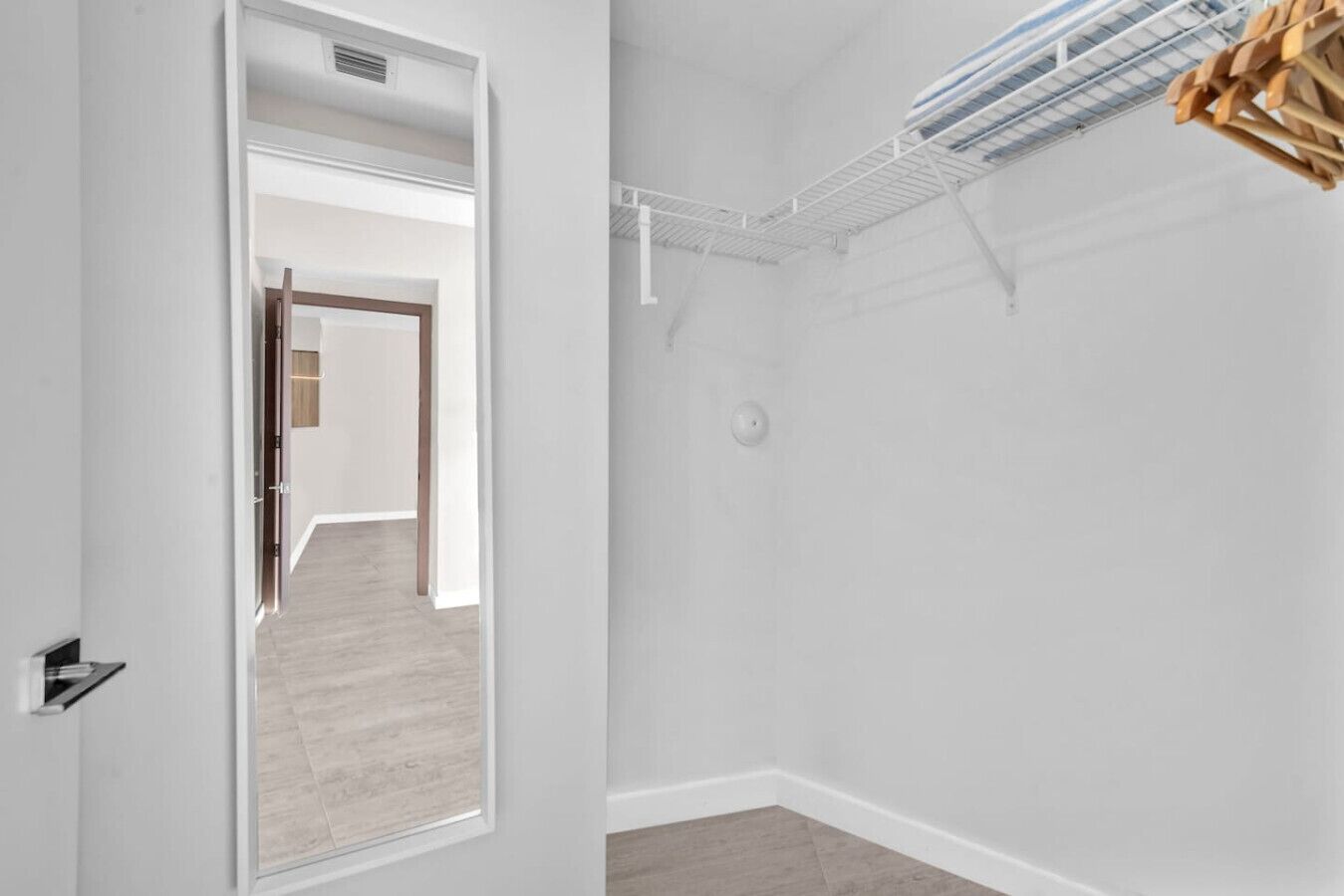A room with a white interior, a wooden floor, a door leading to another room, and a ceiling with a ventilation system.