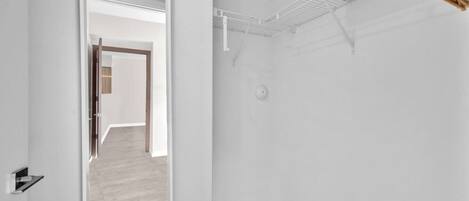 A room with a white interior, a wooden floor, a door leading to another room, and a ceiling with a ventilation system.