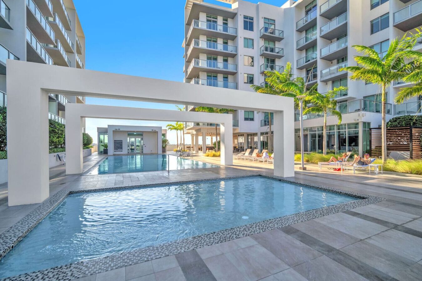 A modern outdoor swimming pool with lounge chairs and palm trees, surrounded by high-rise residential buildings.