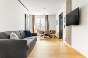 Una sala moderna con un sofá gris, un televisor de pantalla plana montado en la pared, una mesa de comedor con sillas y piso de madera.