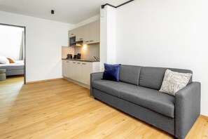 A modern living room with a grey sofa, a wooden floor, and a kitchen in the background.