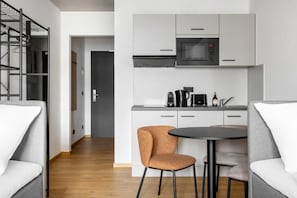 A modern kitchen with a microwave, coffee maker, and cabinets. There is a round table with two chairs and a sofa in the living area.