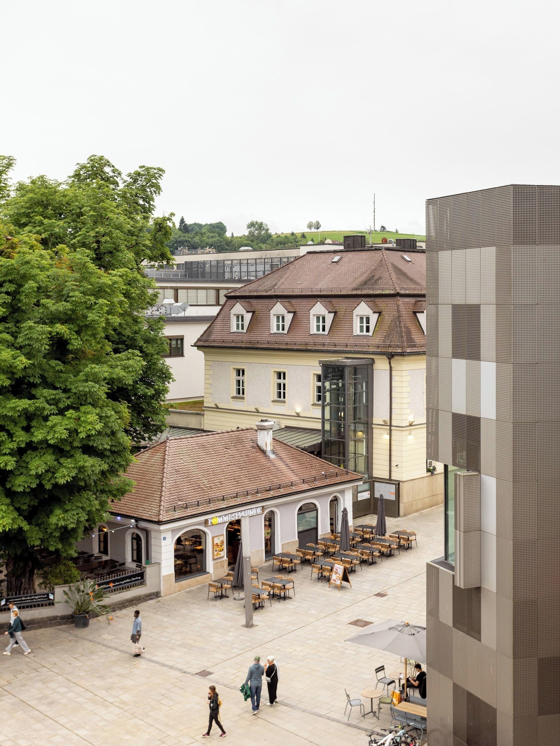 Ein Innenhof mit Außenbestuhlung, ein Gebäude mit Ziegeldach und ein großer Baum.