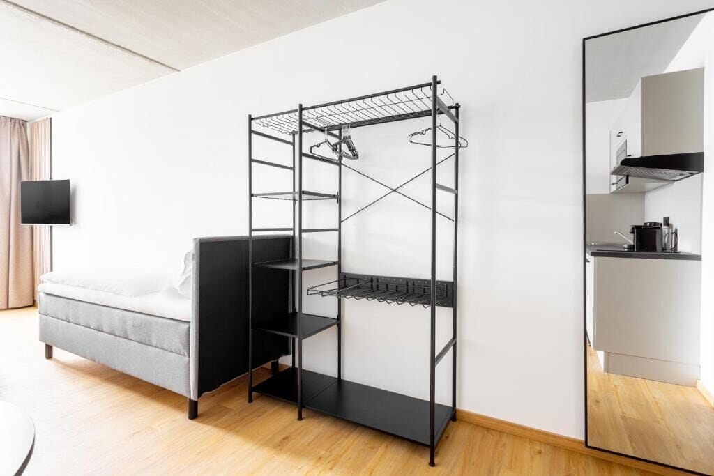 A compact room with a bed, a black metal shelf unit, a flat-screen TV, and a kitchen area.