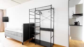 A compact room with a bed, a black metal shelf unit, a flat-screen TV, and a kitchen area.