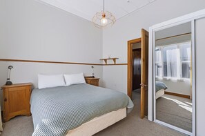 A bedroom with a bed, bedside lamps, a wooden nightstand, a mirror, and a door leading to another room. - Hannan House - Whitianga Holiday Home (Whitianga)