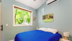 Un dormitorio con un ventanal grande, una cama con cubrecama azul, una mesita de noche con lámpara y un aire acondicionado instalado en la pared.