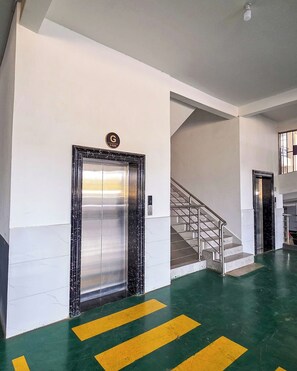 Un espace intérieur doté d’un ascenseur métallique, d’un escalier et d’un sol marqué de motifs jaunes et verts.