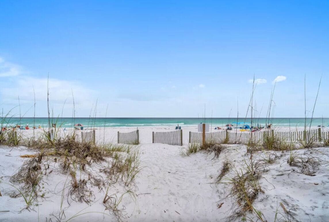 A beach with a wooden fence leading to the water, sandy beach, and a clear sky.